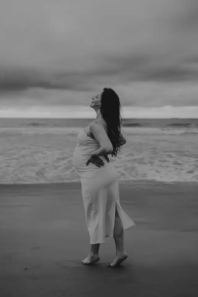 Gestante de perfil na praia, vestido claro, mãos na cintura