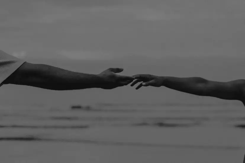 Dois braços se aproximando sobre a praia, pontas dos dedos quase se tocando